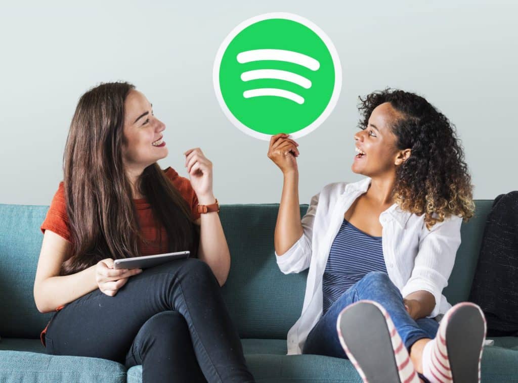A imagem mostra uma mulher branca e uma mulher negra sentadas em um sofá. Uma delas, está segurando o logotipo do Spotify