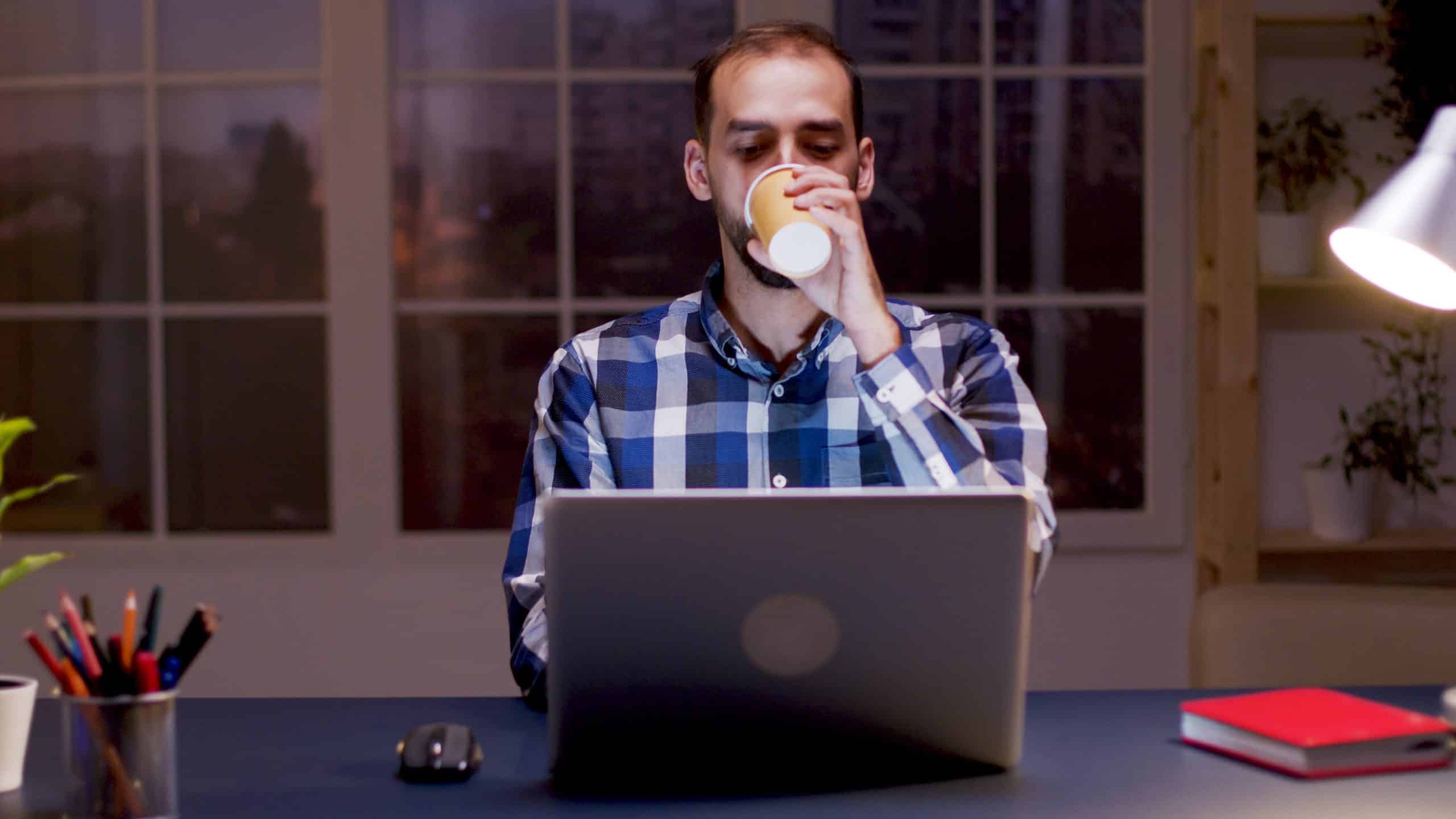 A imagem mostra um homem branco, desenvolvedor remoto, tomando café enquanto trabalha sentado à frente de um notebook