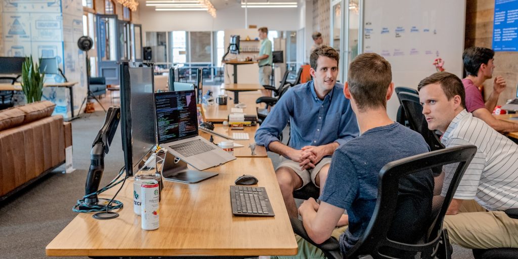 A imagem representa o ambiente corporativo de uma empresa de outsourcing, com três homens brancos sentados conversando em um escritório