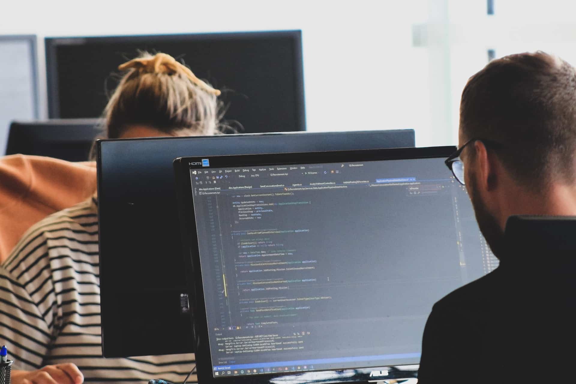 A imagem mostra uma mulher e um homem à frente de dois computadores, trabalhando como desenvolvedores back-end