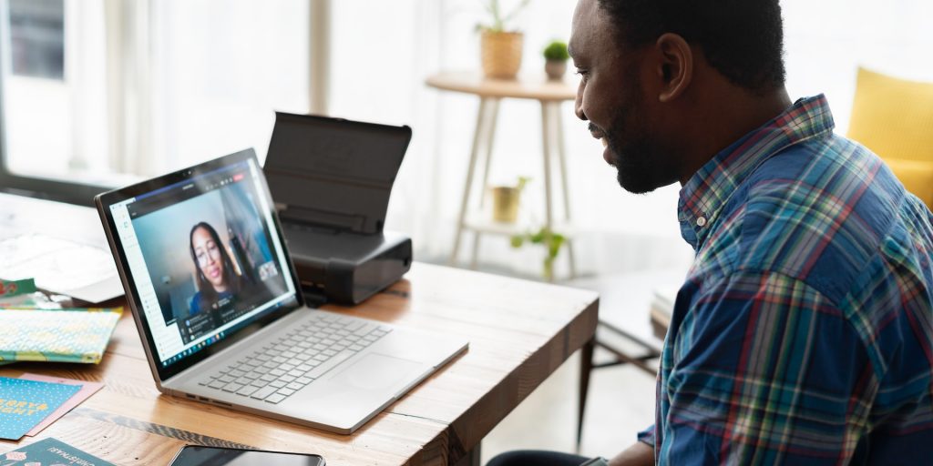 A imagem mostra um homem negro conversando com uma mulher negra através de uma vídeo-conferência, representando uma entrevista para uma vaga de desenvolvedor remoto